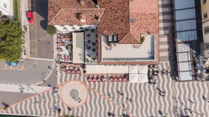 Vista aérea de una zona costera con una plaza empaveda, mesas al aire libre y un edificio de tejado rojo.