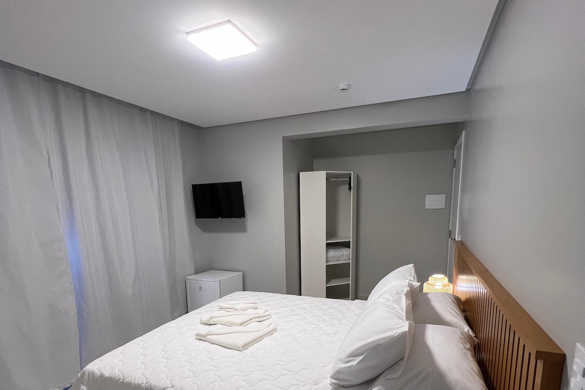 A neatly made bed with white linens, a bedside table, a wardrobe, and a flat-screen TV mounted on the wall.