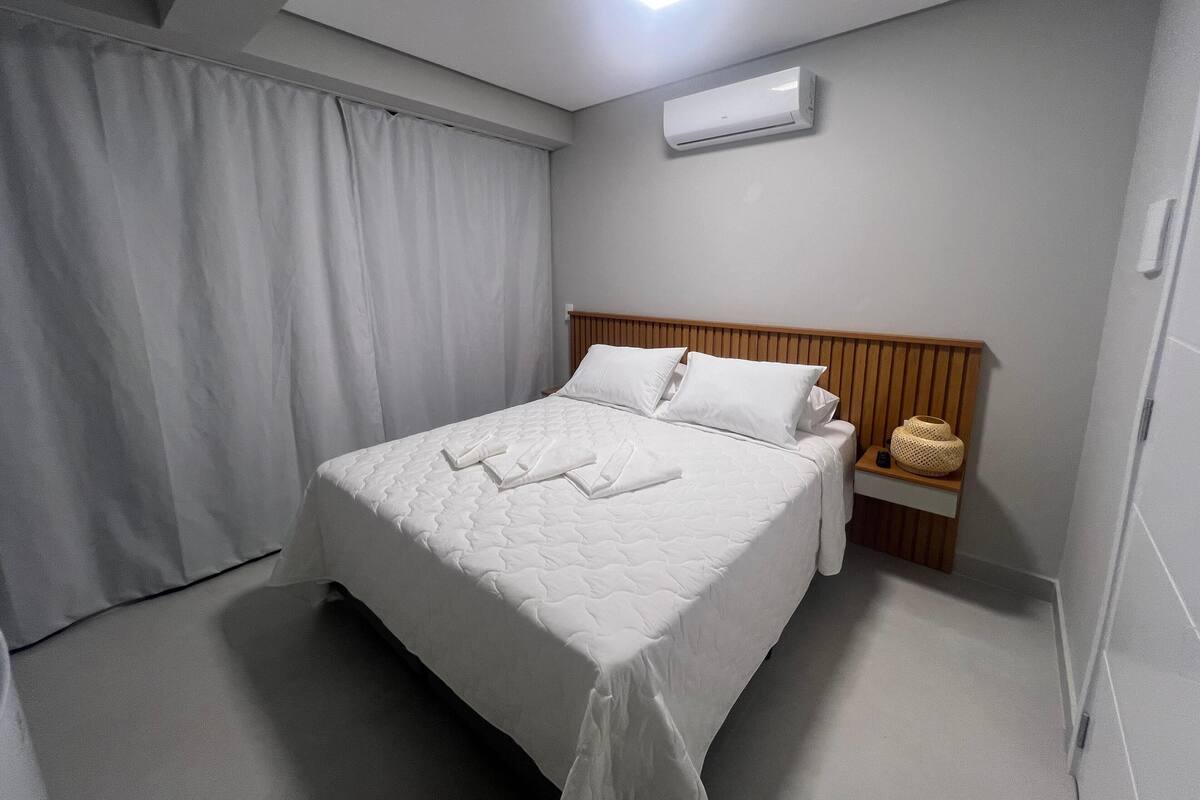 A neatly made bed with a wooden headboard, a bedside table with a lamp and a basket, and an air conditioning unit on the wall.