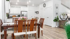 Una cocina moderna con armarios blancos, un refrigerador de acero inoxidable y un comedor con sillas y mesa de madera.