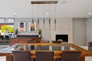 A modern dining area with a glass-top table, brown chairs, pendant lights, and a fireplace. - Private Modern Home w Pool, Firepit & BBQ - Enjoy AZ outdoor living at its best! (Scottsdale)