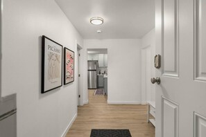 A hallway with white walls, a wooden floor, and a kitchen visible in the distance. - 2BR Lux Apartment | 5 Mins from Airport | Pets+ (Burien)