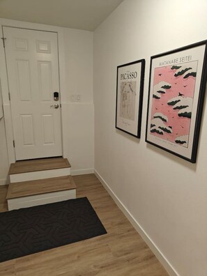 A hallway with a white door, a staircase with a black doormat, and two framed artworks on the wall. - 2BR Lux Apartment | 5 Mins from Airport | Pets+ (Burien)