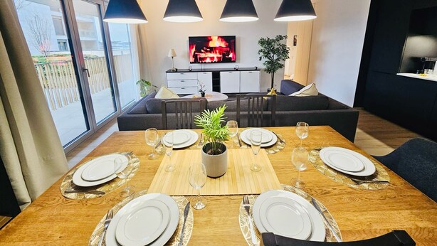 Un comedor moderno con una mesa de madera dispuesta para seis personas, lámparas colgantes negras y un televisor al fondo.