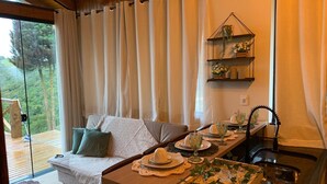 Une pièce chaleureuse avec un canapé, une table à manger dressée avec des assiettes et des verres, une étagère suspendue ornée de fleurs et une vue sur une forêt, visible à travers une porte vitrée.