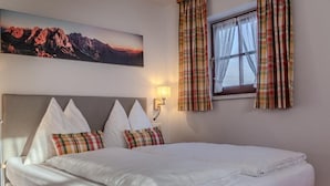 A neatly made bed with white linens and decorative pillows, a wall-mounted lamp, a window with plaid curtains, and a mountain landscape painting above the bed.