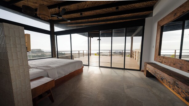Een slaapkamer met een groot bed, een houten bankje en een balkon met uitzicht op het strand.