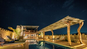 Een zwembadgedeelte bij nacht met ligbedden, een zwembad en een houten pergola.