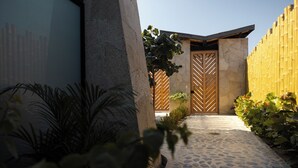 Een pad van stenen dat leidt naar een houten poort met een geometrisch ontwerp, omringd door planten en een bamboe hek.