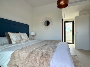 A bedroom with a large bed, a round mirror, a lamp, and a door leading to an outdoor area.