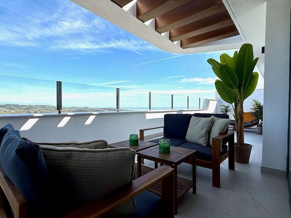 A rooftop terrace with a view of the sea, furnished with a sofa, a table, and a potted plant.