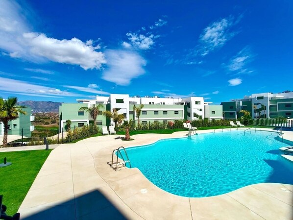 Ein Swimmingpool mit blauem Himmel, moderner Architektur im Hintergrund und einem gepflegten Rasen.