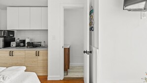 A compact kitchen with white cabinets, a microwave, and a coffee maker. A wooden cabinet and a small wooden table are visible in the background. - Charming Studio Dennis Port Getaway with WiFi and AC - Close to Beach! (Dennis)