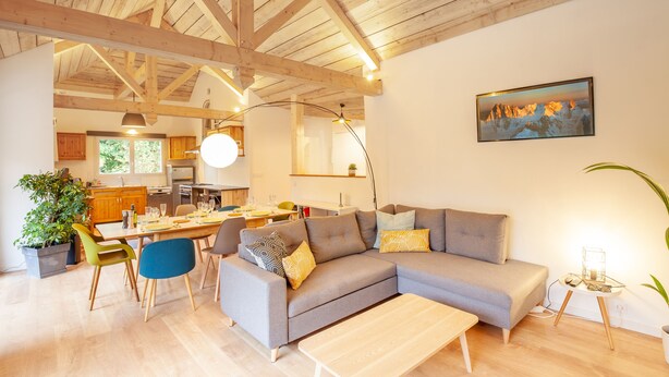 A modern living room with a grey sectional sofa, wooden dining table, and a kitchen in the background.