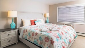 Un dormitorio con cama, mesitas de noche, lámparas, una ventana con persianas y un radiador.