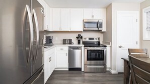 Una cocina moderna con armarios blancos, electrodomésticos de acero inoxidable, microondas, horno, lavavajillas y una mesa de comedor de madera.