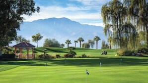 Un campo de golf con un pabellón, palmeras y montañas al fondo.