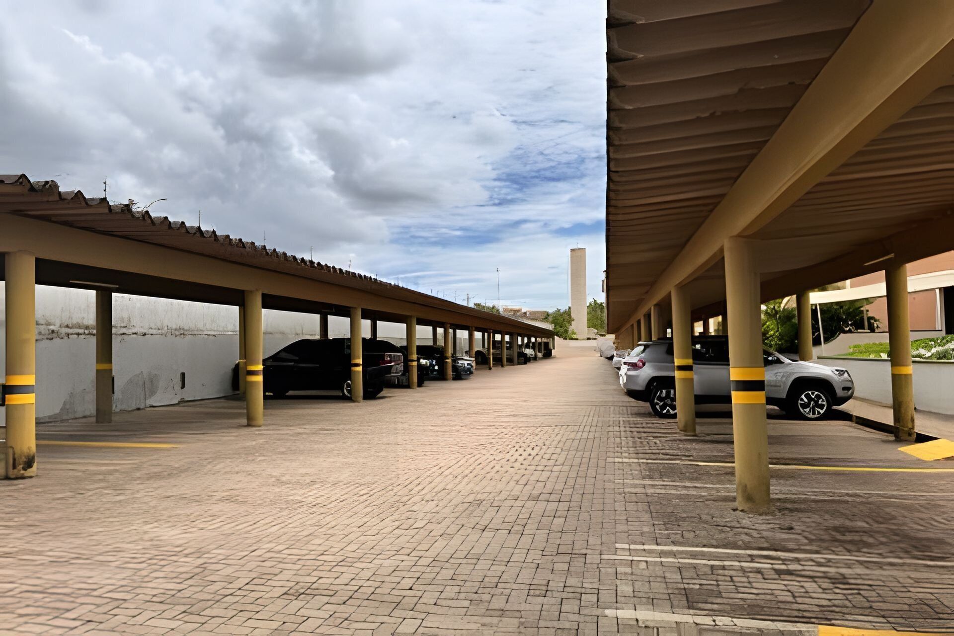 Een overdekte parkeerplaats met meerdere rijen geparkeerde auto's, een bestrate oprit en een hoge constructie op de achtergrond.