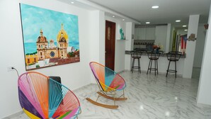 A modern interior with two colorful rocking chairs, a large painting of a coastal church, and a kitchen area with bar stools. - Casa Sandunguera (Cartagena)