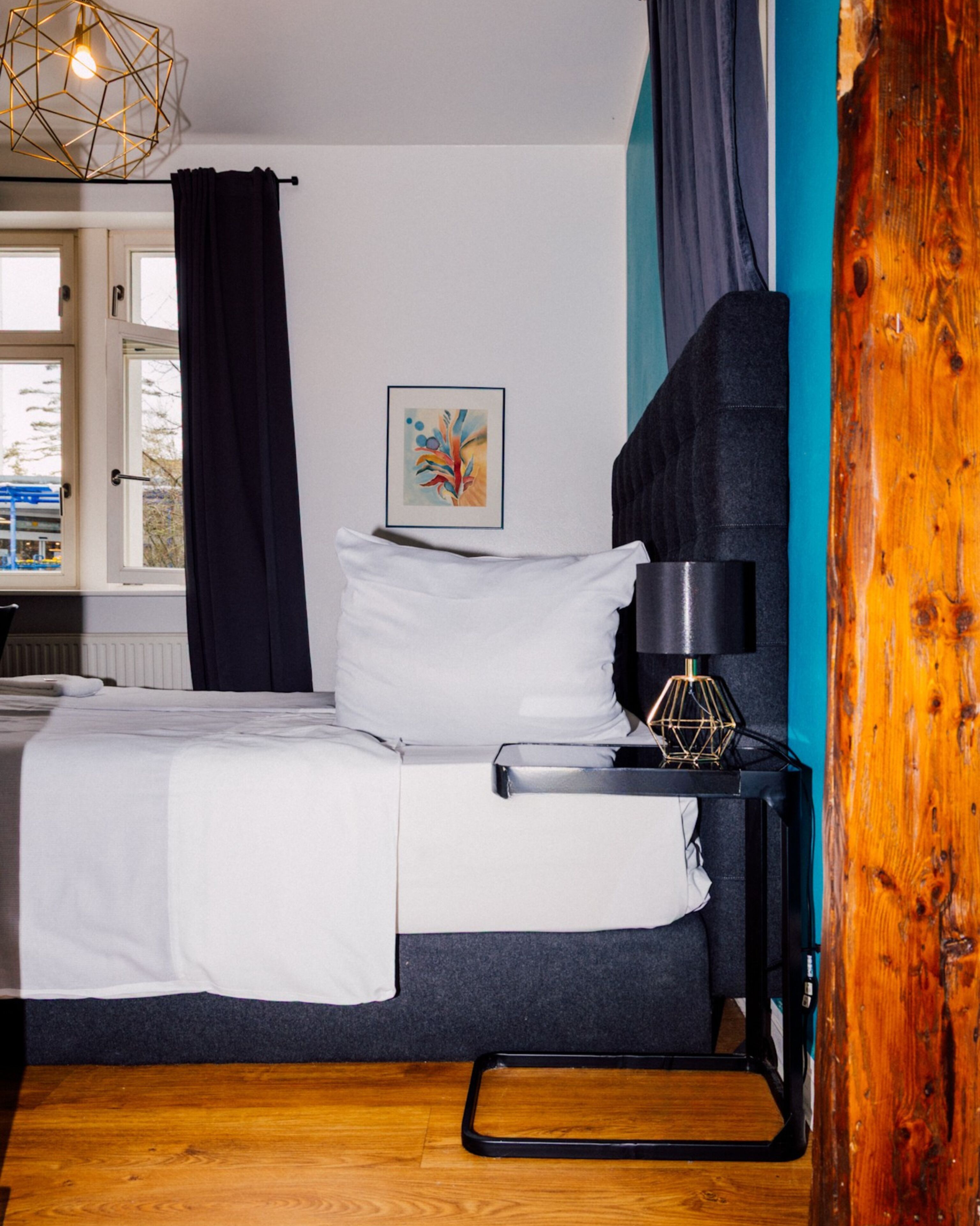 Une chambre à coucher avec un lit, une table de chevet avec une lampe, une fenêtre avec des rideaux et un tableau au mur.