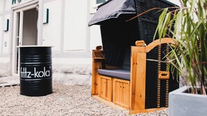 Een houten terras met buitenmeubels, een zwart zonnescherm en een zwarte ton met de tekst 'fritz-kola', met een hotelschilt in de verte.