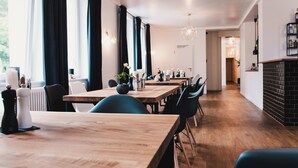 Een moderne eetruimte met houten tafels en stoelen, met een keuken in de achtergrond en een gang die naar andere delen van het gebouw leidt.