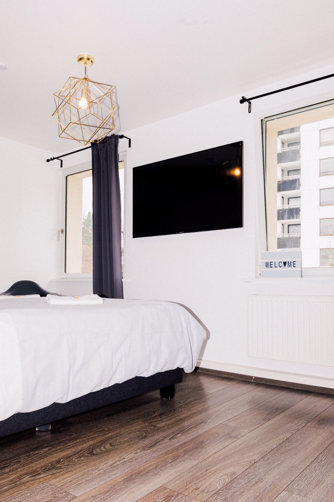 A modern hotel room with a bed, a TV mounted on the wall, a window with a view of buildings, and a unique geometric light fixture.