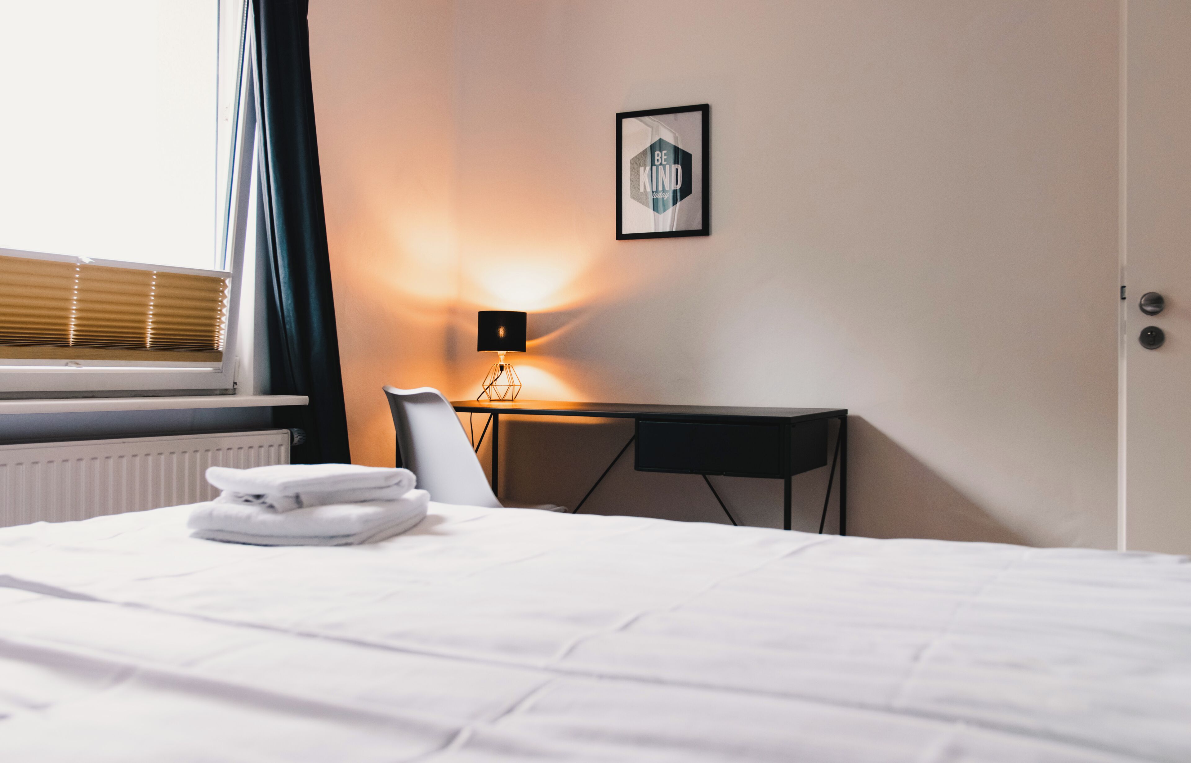 A hotel room with a bed, a desk, a chair, a radiator, a window with blinds, and a wall-mounted picture.