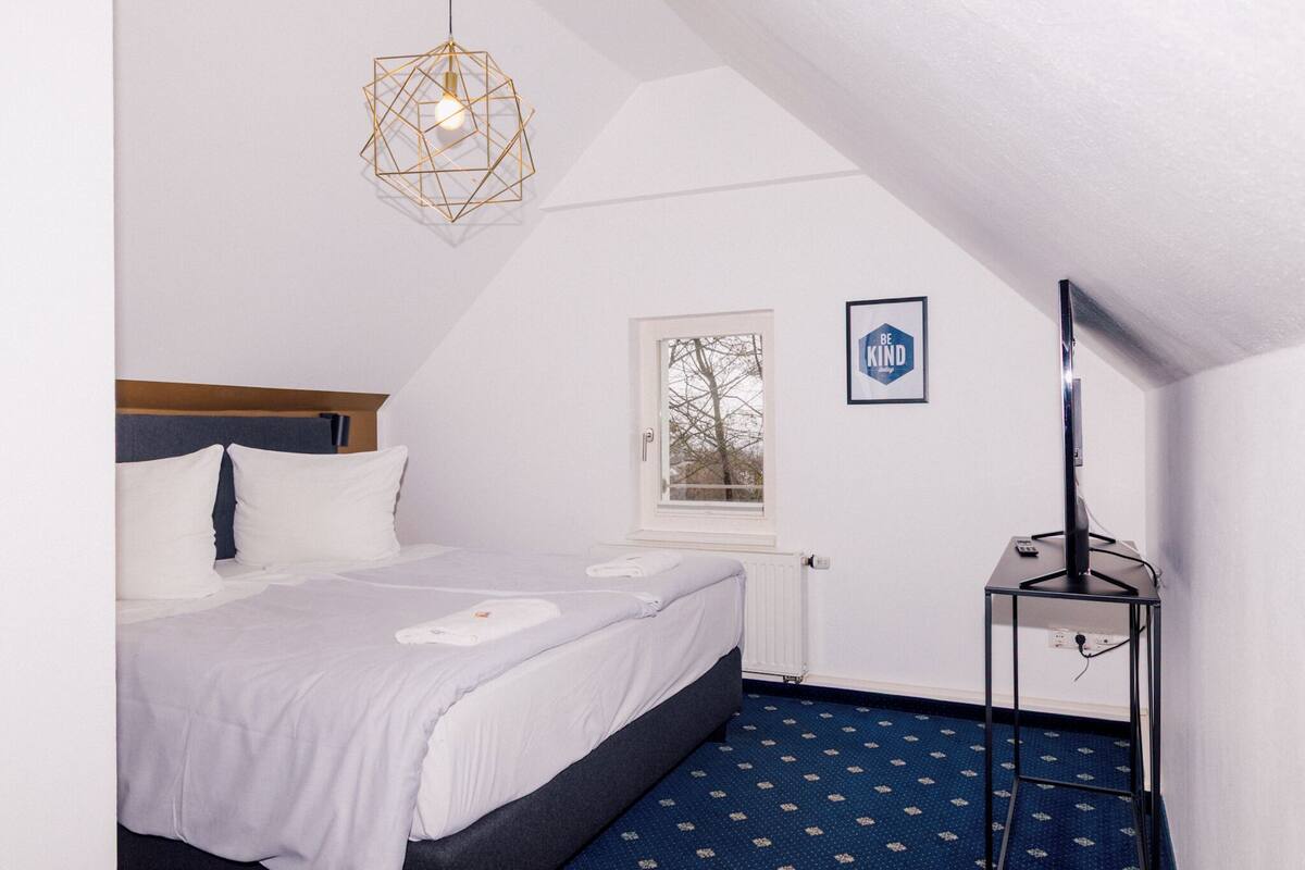 Ein Schlafzimmer mit einem Bett, einem Fenster, einem an der Wand montierten Fernseher und einer geometrischen Leuchte.
