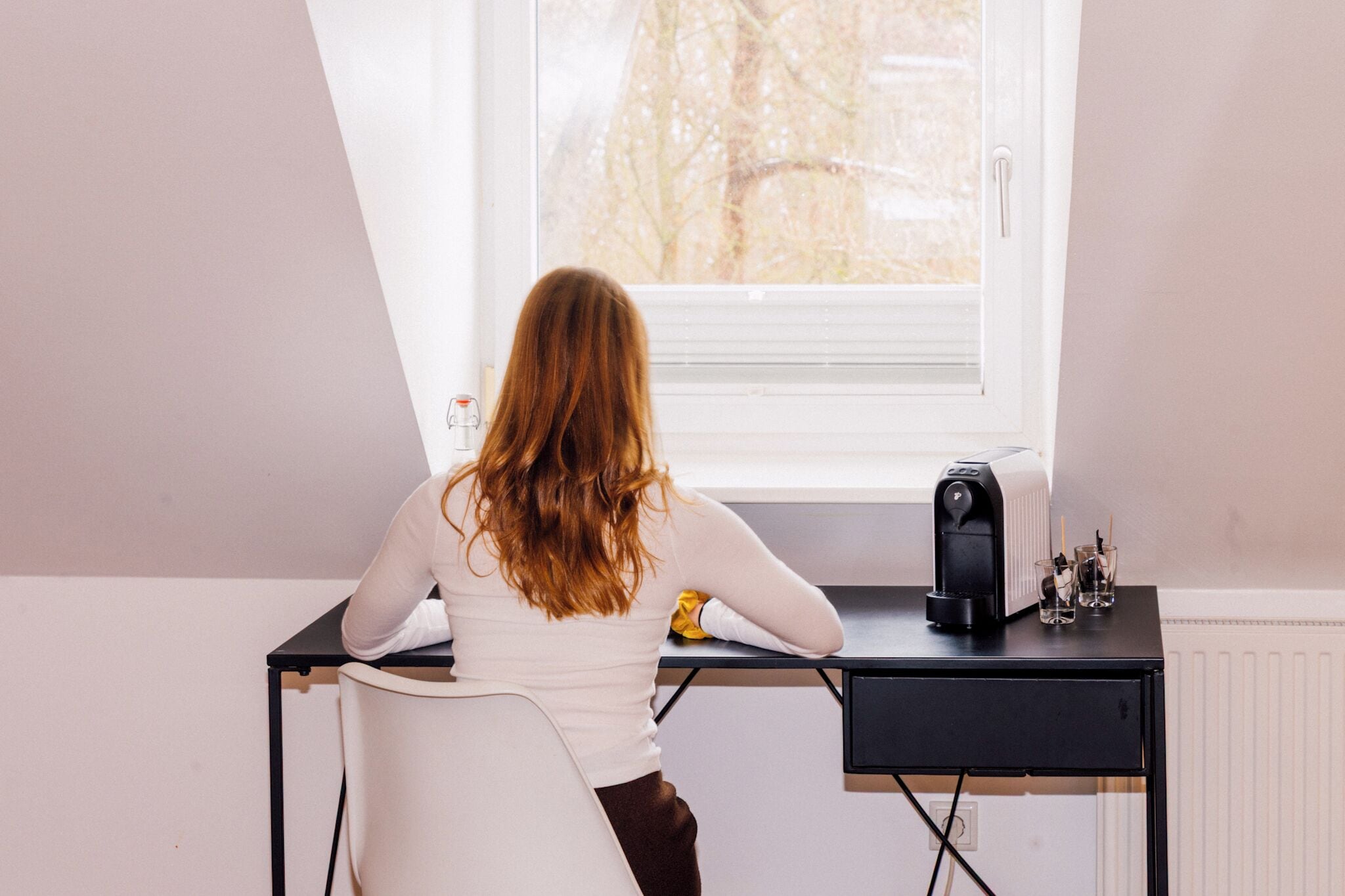 A person seated at a desk with a window, a radiator, and a coffee maker on the table.
