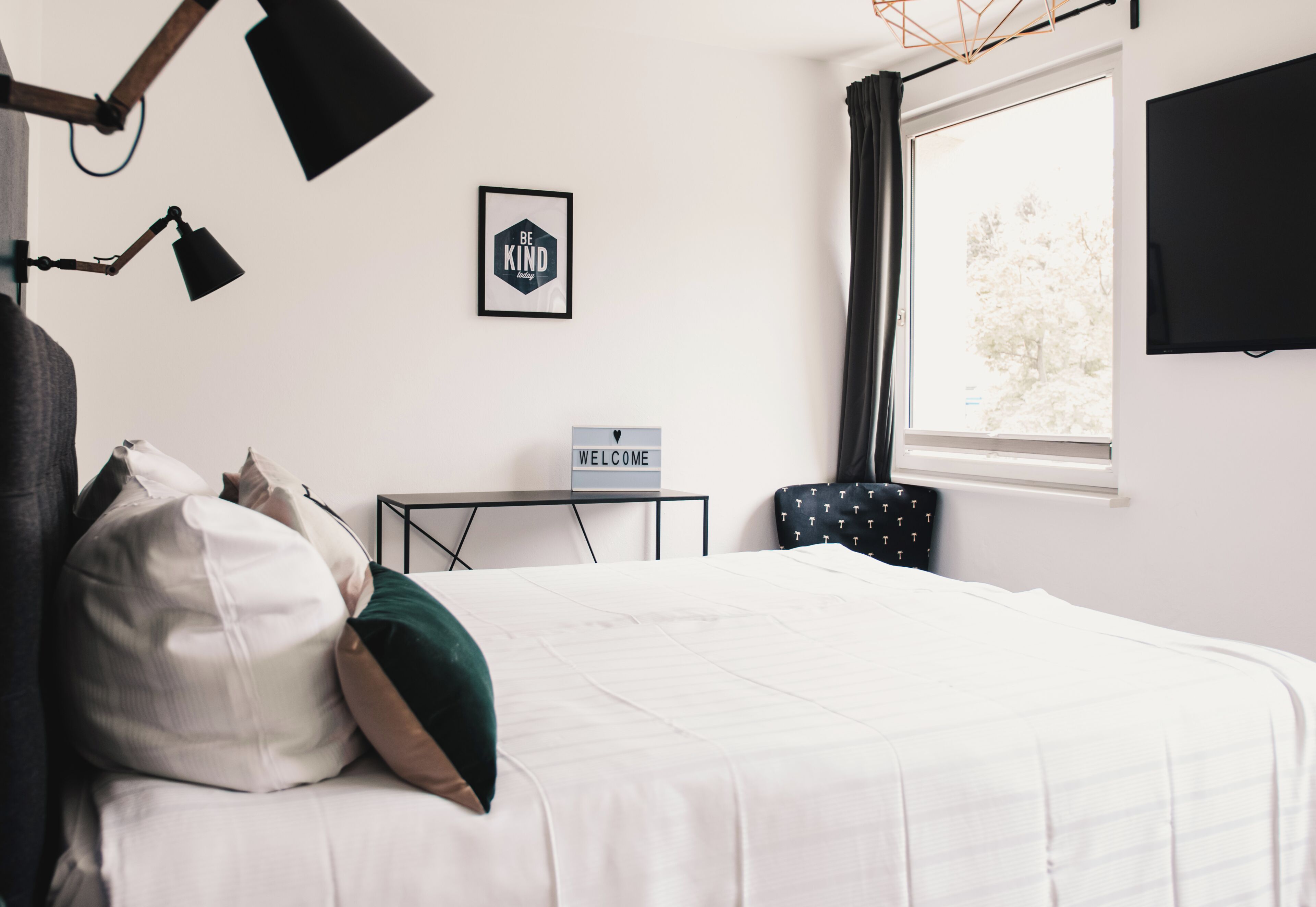 Hotelkamer met een bed, een 'WELCOME' bord, een bureau, een stoel, een televisie en een raam met gordijnen.