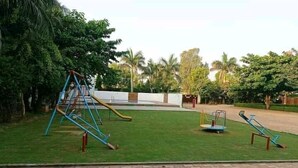 Un parque infantil con columpios y tobogán, rodeado de follaje y una zona pavimentada.