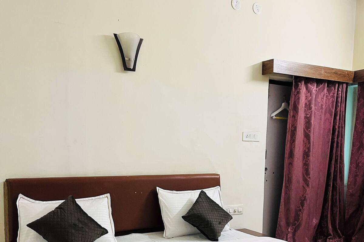 Quarto de hotel com cama, luminária na parede, cabideiro e porta com cortinas.