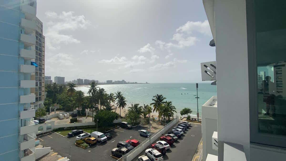 A view of a beach with palm trees, a parking lot with cars, and a high-rise building in the background.