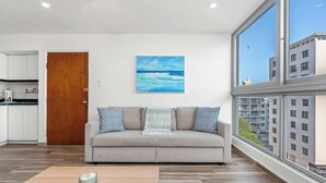 Ein modernes Wohnzimmer mit einer Couch, einem Gemälde des Meeres und Blick auf Gebäude durch große Fenster.