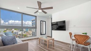 Ein modernes Wohnzimmer mit großem Fenster, Deckenventilator, Flachbildfernseher, Couchtisch aus Holz und Korbstühlen.