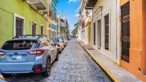 Una estrecha calle adoquinada flanqueada por edificios coloridos, coches estacionados y un cielo azul despejado.