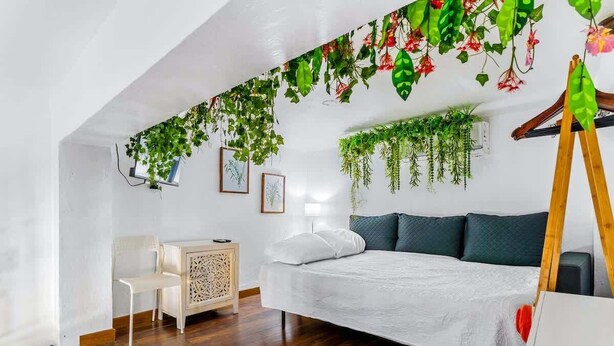 Un dormitorio con techo inclinado, suelo de madera, una cama con ropa blanca, una silla blanca, una mesita de noche de madera y plantas colgantes.