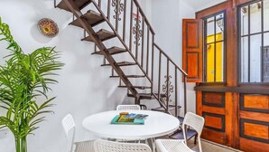 Comedor con una mesa redonda, sillas blancas, una planta en maceta y una escalera con barandilla ornamentada.