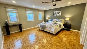 A bedroom with a wooden parquet floor, a bed with a white and blue floral bedspread, two bedside tables with lamps, and three windows with blinds. - Luxury Modern Retreat: w/ Sauna & Modern Amenities (Mississauga)