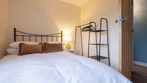 A neatly made bed with white linens and two brown pillows, a bedside lamp, and a metal shelf unit with clothes hanging on it. - 3br-garden-freeparking-neartowncentre (Ipswich)
