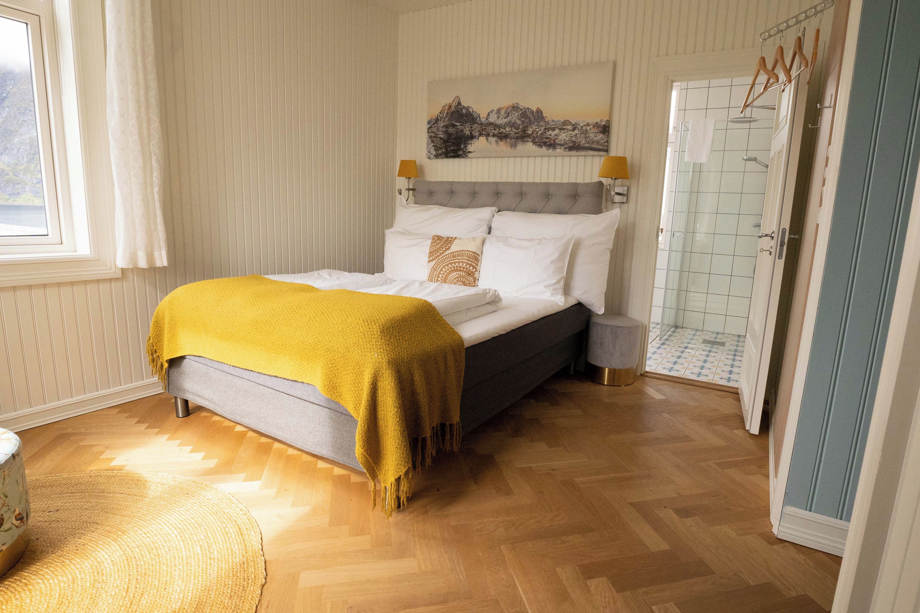 A bedroom with a bed, a bedside table, a lamp, a window, and a door leading to a bathroom.