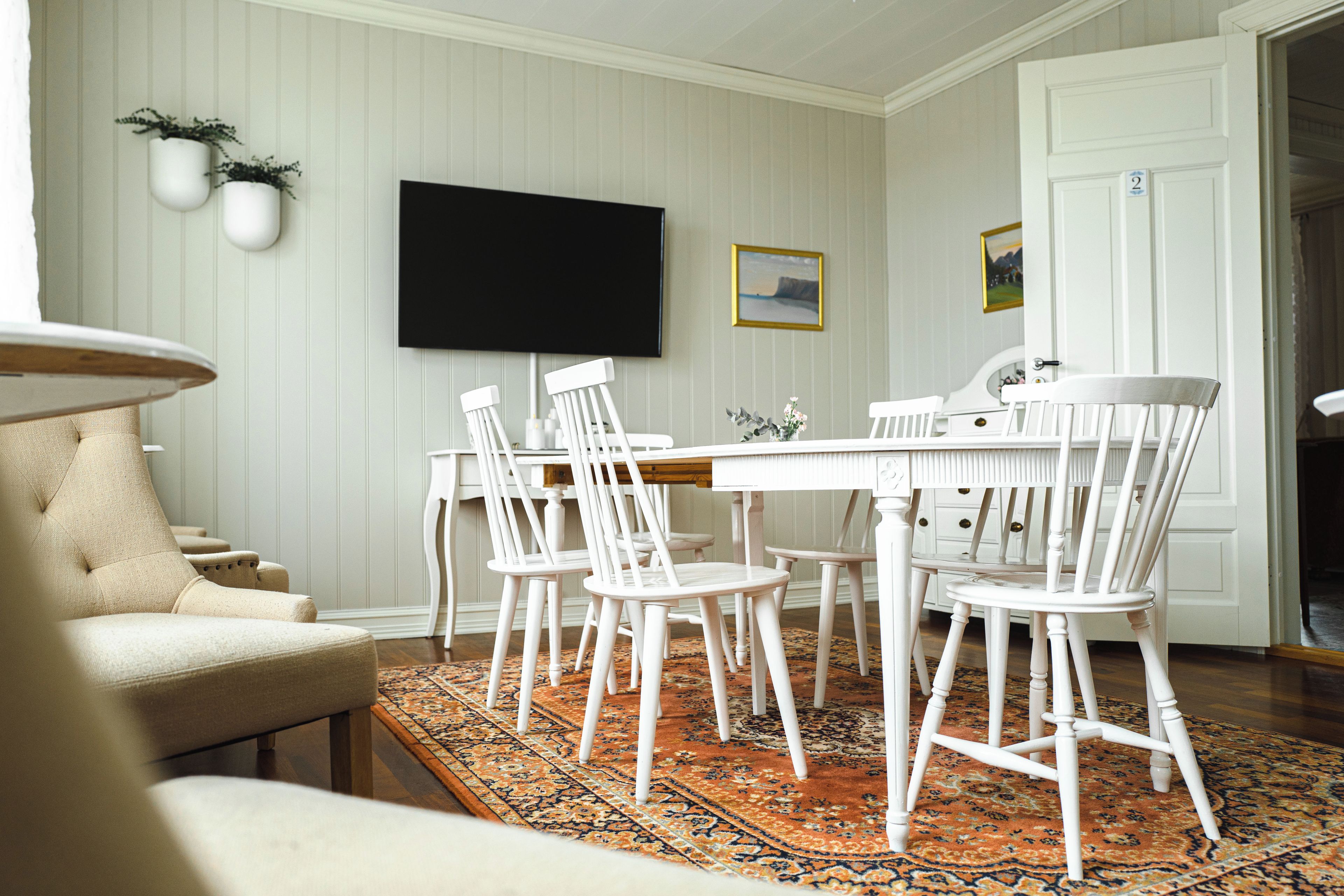 A dining area with a white table and chairs, a flat-screen TV mounted on the wall, and a patterned rug on the floor.