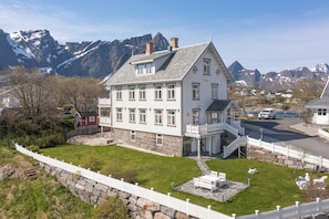 A large white building with multiple windows and a stone foundation, surrounded by a white picket fence and a stone wall. - Sakrisøy Gjestegård-Solbakken anno 1880 (Nordland)