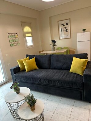 Un salon avec un canapé foncé, deux petites tables rondes ornées de plantes, un coin repas avec une table et des chaises, et un réfrigérateur.