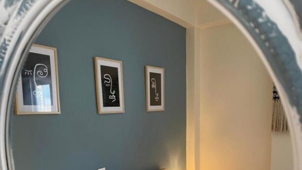 Ein Bett mit weißer Bettwäsche und zwei blauen Kissen, ein Nachttisch mit einem Glas und einem Buch sowie drei gerahmte Kunstwerke an der Wand.