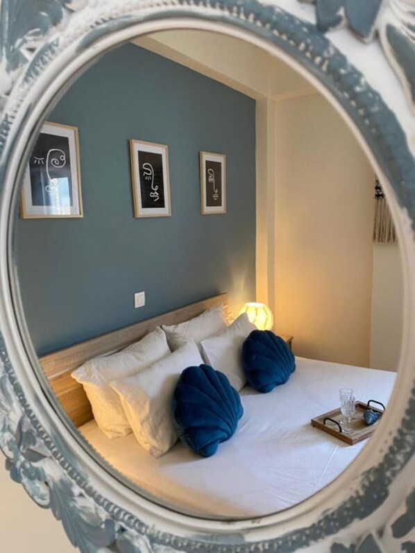 A bed with white bedding and two blue pillows, a bedside table with a glass and a book, and three framed artworks on the wall.