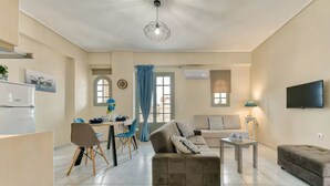 Ein modernes Wohnzimmer mit Sofa, Hocker, Essbereich mit Stühlen, einem Fernseher und einer Deckenleuchte.