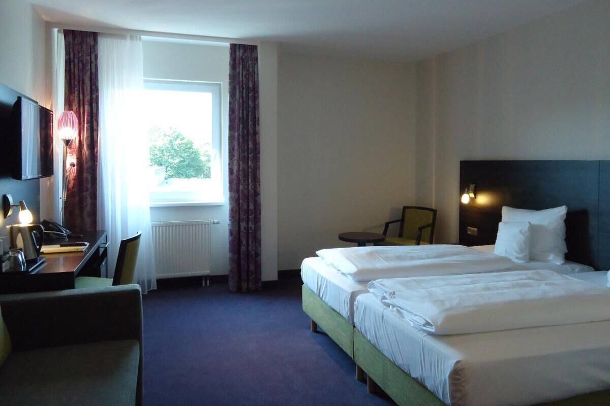 A hotel room with two beds, a desk with a lamp, a chair, and a view of the outdoors through a window.