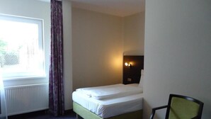 Una habitación de hotel pequeña y ordenada, con una cama individual, una silla, una mesita de noche y un ventanal con cortinas.